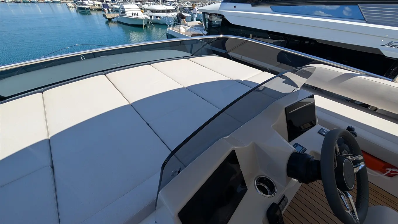 A luxury boat's helm with a steering wheel, control panel, and plush seating overlooking a marina filled with yachts.